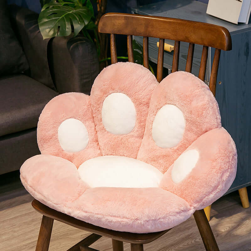 Plush cat paw shaped seat cushion in pink, placed on a wooden chair in a kids' room setting.